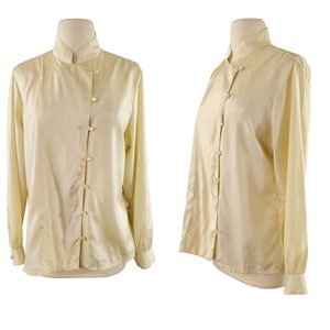 Pode incluir: Uma blusa de manga comprida de cor creme com colarinho mandarim e fecho de botões. A blusa é feita de um tecido sedoso e tem um caimento solto.