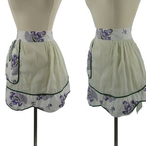 Puede incluir: Un delantal blanco con un borde a cuadros verde y blanco y estampado floral morado. El delantal tiene un solo bolsillo en el lado izquierdo.