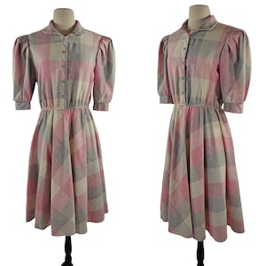 Könnte beinhalten: Ein rosa-graues kariertes Kleid mit Knopfleiste und Puffärmeln. Das Kleid hat ein tailliertes Oberteil und einen weiten Rock.