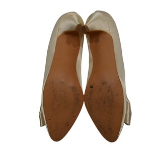 May include: A pair of ivory satin pumps with a pointed toe and a low heel. The shoes have a bow detail on the back.