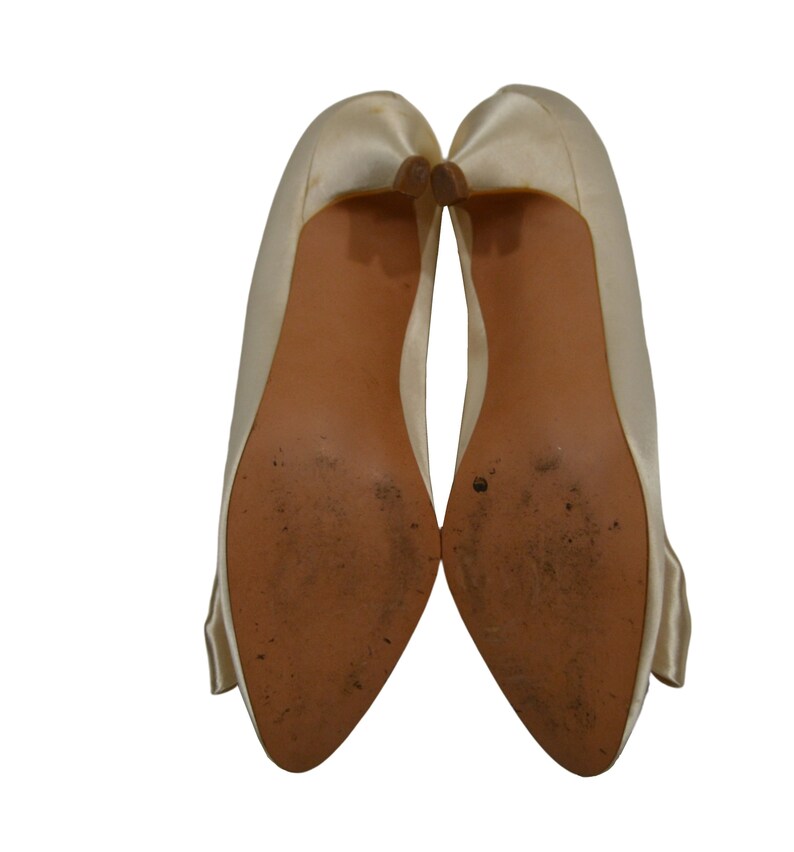 May include: A pair of ivory satin pumps with a pointed toe and a low heel. The shoes have a bow detail on the back.