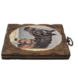 May include: A wooden coaster with a painted image of two horses. The horses are brown and black, with one wearing a bridle. The coaster has a dark brown, textured edge and a metal ring for hanging.