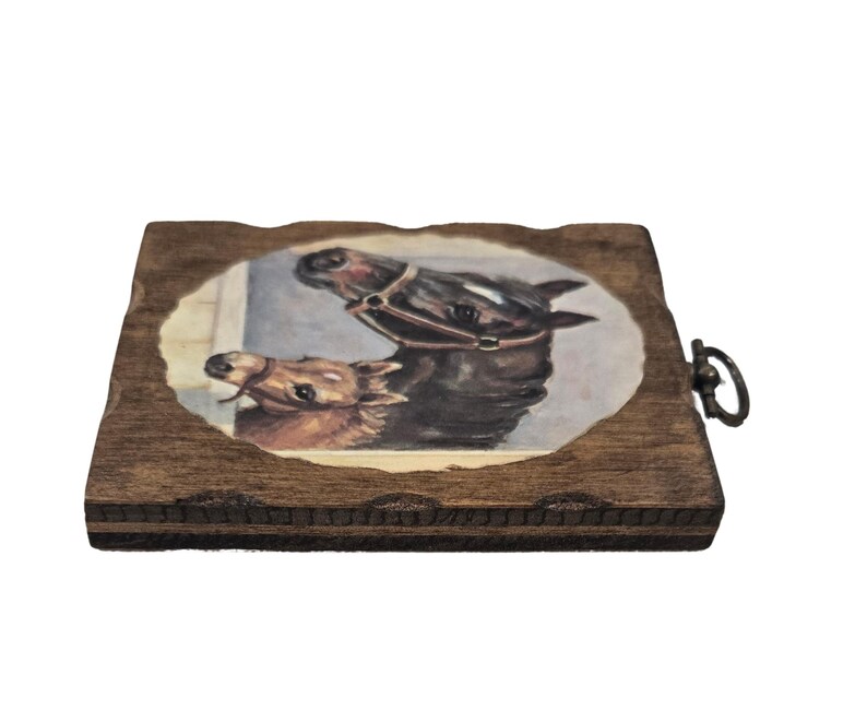 May include: A wooden coaster with a painted image of two horses. The horses are brown and black, with one wearing a bridle. The coaster has a dark brown, textured edge and a metal ring for hanging.