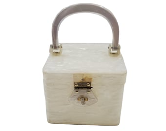 May include: A white, square-shaped handbag with a white plastic handle and a gold clasp.