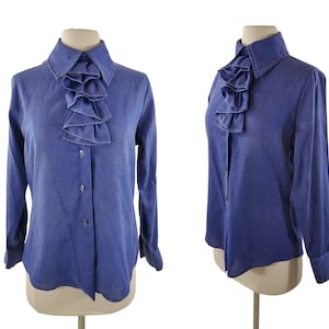 May include: A blue denim shirt with a large ruffled collar. The shirt has a button-up front and long sleeves.