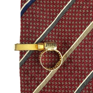 Puede incluir: Pinza de corbata dorada con un anillo y el número 6202 grabado en él. La pinza de corbata está en una corbata a rayas rojas, blancas y azules.