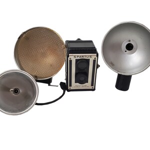 May include: A black vintage Spartus Full-Vue camera with two flash attachments. One flash attachment is a silver reflector with a black handle, and the other is a silver reflector with a black handle and a yellow diffuser.