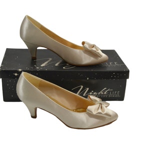 May include: A pair of ivory satin pumps with a small heel and a satin bow on the toe. The shoes are on a black box with the text "Night Life by Life Brides".