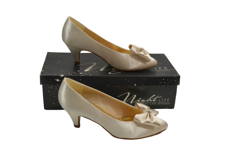 May include: A pair of ivory satin pumps with a small heel and a satin bow on the toe. The shoes are on a black box with the text "Night Life by Life Brides".