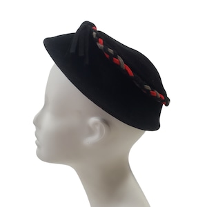 May include: A black velvet hat with a braided trim in red, black, and beige. The hat has a small tassel.