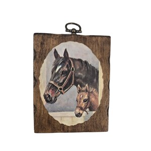 May include: A framed horse portrait. The artwork features a dark brown horse and a light brown foal, both wearing bridles. The painting is set within an oval shape, framed by a dark wooden plaque with a metal loop for hanging.
