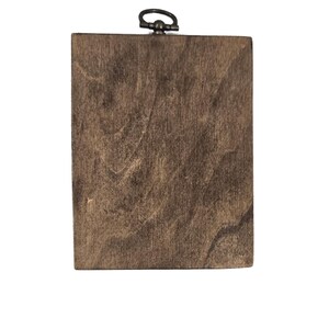 May include: A rectangular wooden plaque with a dark brown finish. The wood grain is visible, creating a natural texture. A small, dark metal ring is attached to the top for hanging. The plaque is set against a white background.
