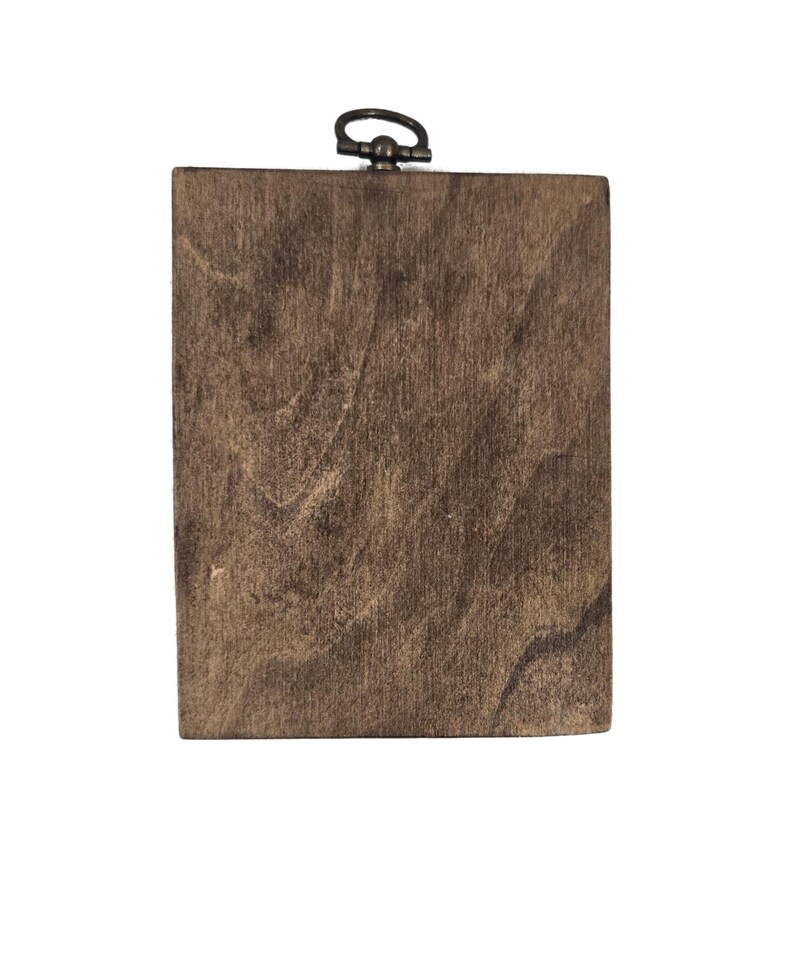 May include: A rectangular wooden plaque with a dark brown finish. The wood grain is visible, creating a natural texture. A small, dark metal ring is attached to the top for hanging. The plaque is set against a white background.