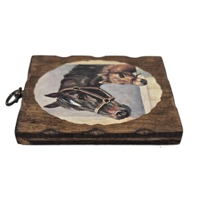 May include: A wooden plaque with a painted oval image of two horses. The horses are brown and black, with one wearing a bridle. The plaque has a dark brown finish and a metal ring for hanging.