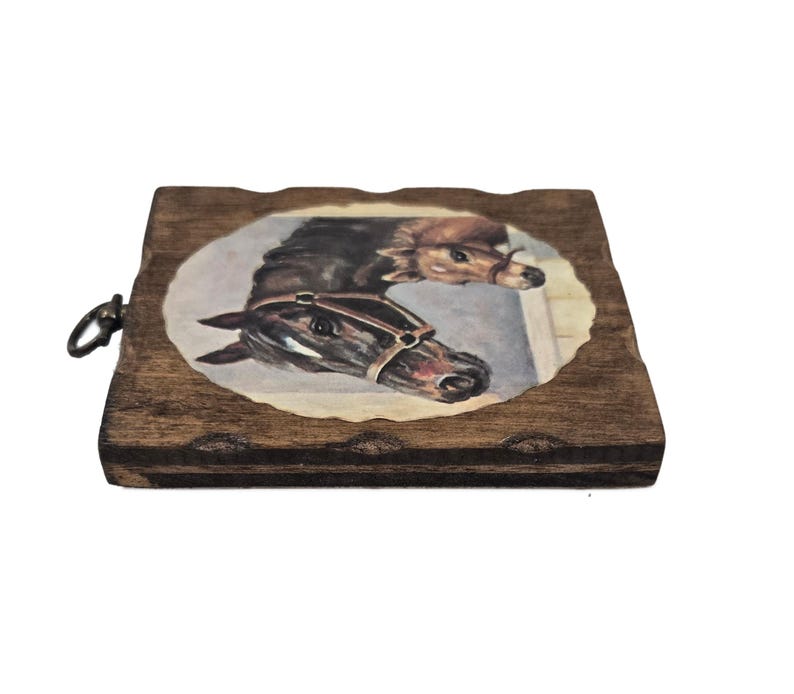 May include: A wooden plaque with a painted oval image of two horses. The horses are brown and black, with one wearing a bridle. The plaque has a dark brown finish and a metal ring for hanging.