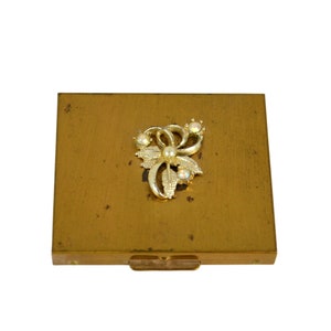 May include: A gold metal compact with a decorative floral design featuring a pearl and rhinestones.