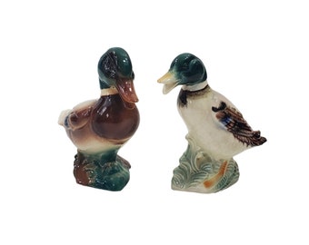 May include: Two ceramic duck figurines. One duck is brown and white with a green head and the other duck is white with a green head and brown and blue markings. Both ducks are standing on a green base.