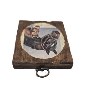 May include: A small, wooden box with a dark stain and a brass-colored handle. The lid features a painted image of two horses, one brown and one dark brown, wearing a harness. The image is set within an oval frame.