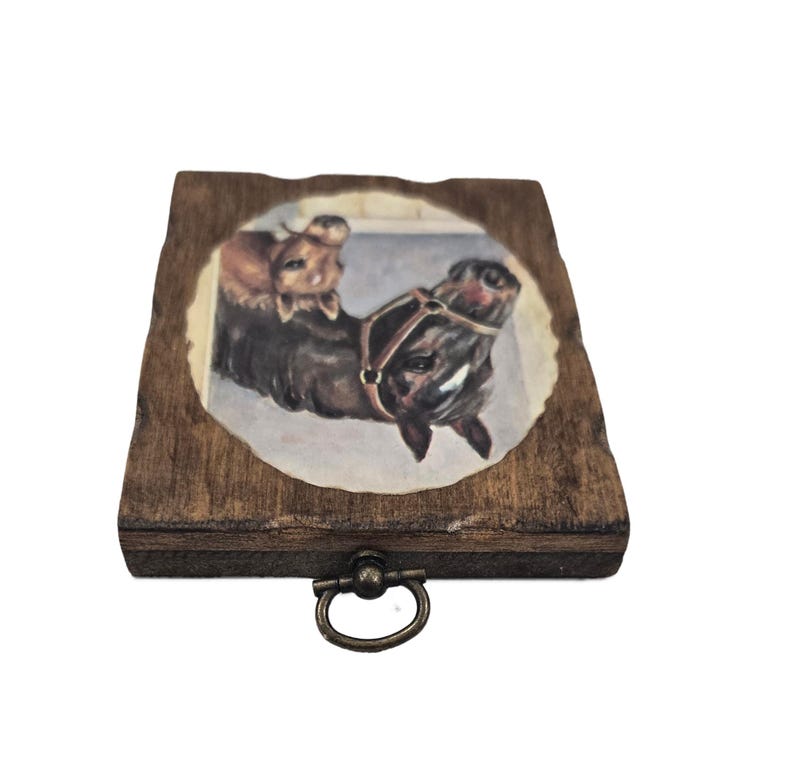 May include: A small, wooden box with a dark stain and a brass-colored handle. The lid features a painted image of two horses, one brown and one dark brown, wearing a harness. The image is set within an oval frame.