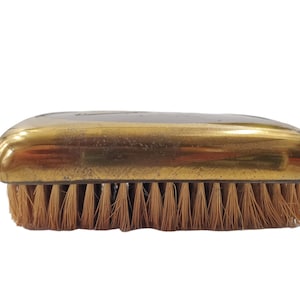 May include: A gold-colored brush with a wooden handle and natural bristles. The brush is designed for cleaning and grooming.