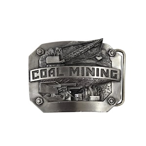 May include: A rectangular silver-toned metal belt buckle featuring a raised relief design of a coal mining scene. The top shows a large crane, while the bottom depicts miners in a tunnel. The text COAL MINING is embossed across the center.
