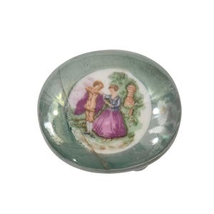 May include: A small round porcelain dish with a metallic green border and a central illustration of two figures in period clothing standing in a garden setting. The dish features a visible crack running across the left side of the surface.