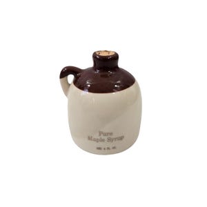 Puede incluir: Una pequeña jarra de cerámica con asa, con la parte superior y el asa marrones, y una base de color crema. La jarra lleva la etiqueta "Pure Maple Syrup" y está diseñada para contener 118 ml de jarabe.
