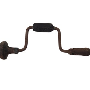 Puede incluir: Un taladro manual antiguo con un mango y pomo de madera oscura. El marco metálico del taladro es de color marrón óxido y el mango central es negro. Esta herramienta vintage está diseñada para taladrar manualmente.