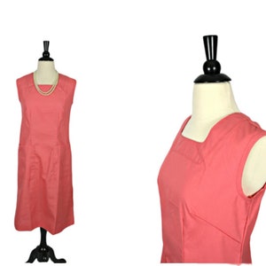 Könnte beinhalten: Ein ärmelloses korallenfarbenes Kleid mit quadratischem Ausschnitt. Das Kleid befindet sich auf einer Schaufensterpuppe und hat ein schlichtes, klassisches Design.