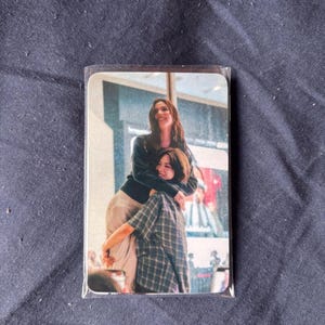 May include: A rectangular photo card encased in a clear plastic sleeve featuring a color image of two individuals embracing. One person with long dark hair wears a black long-sleeved top and light trousers, while the other wears a plaid shirt.