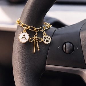 May include: Gold-toned charm bracelet with three dangling charms: a white circle with the letter "A", a bow, and a paw print. The bracelet is hanging on a black steering wheel.