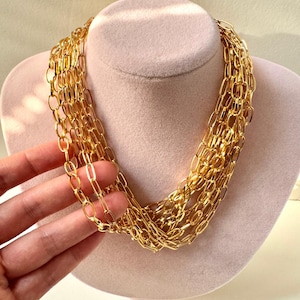 May include: A multi-strand gold chain necklace featuring elongated oval links. The necklace consists of several layers of polished gold-toned metal chains that drape together to create a voluminous, layered appearance around the neck area.