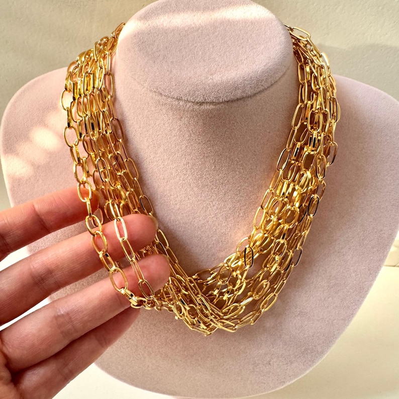 May include: A multi-strand gold chain necklace featuring elongated oval links. The necklace consists of several layers of polished gold-toned metal chains that drape together to create a voluminous, layered appearance around the neck area.