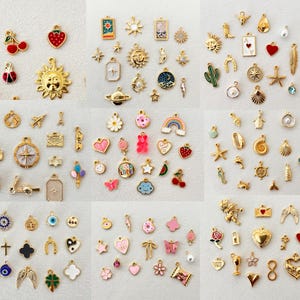 May include: A collection of gold-toned charms in various shapes and designs, including cherries, suns, hearts, and stars. Some charms feature enamel details in red, pink, and blue. The charms are scattered on a white surface.