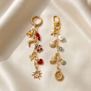 May include: Two gold-toned metal keychains featuring various charms. The left keychain includes a cowboy hat, boot, red chili, cherries, cactus, heart, and sun. The right keychain features a starfish, pearl, shells, blue stone, and spiral shell.