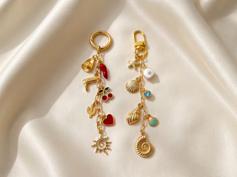 May include: Two gold-toned metal keychains featuring various charms. The left keychain includes a cowboy hat, boot, red chili, cherries, cactus, heart, and sun. The right keychain features a starfish, pearl, shells, blue stone, and spiral shell.