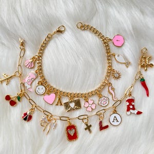 May include: Two gold-tone charm bracelets featuring various enamel and metal charms including a pink heart, a smiley face, a letter A, a red chili pepper, a ladybug, a cowboy boot, a seashell, a paw print, a flower, and a gold-tone airplane.