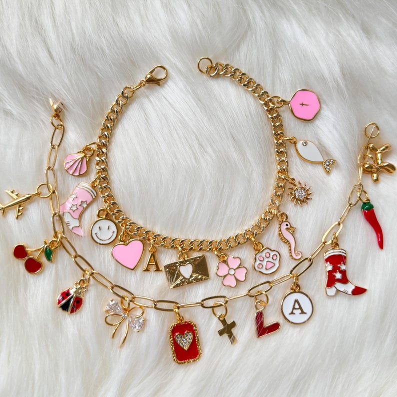 May include: Two gold-tone charm bracelets featuring various enamel and metal charms including a pink heart, a smiley face, a letter A, a red chili pepper, a ladybug, a cowboy boot, a seashell, a paw print, a flower, and a gold-tone airplane.