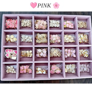 May include: A pink velvet jewelry tray with twenty-four compartments, each containing small metal charms in various shapes like hearts, flowers, shells, and stars. The text PINK is displayed at the top with heart and flower icons.