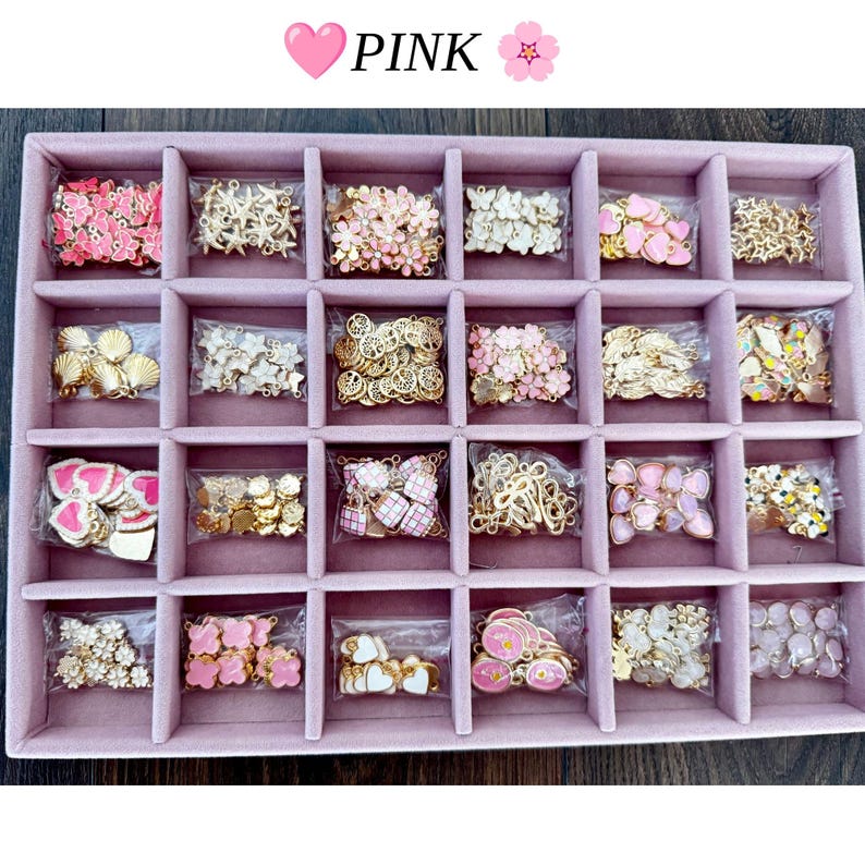 May include: A pink velvet jewelry tray with twenty-four compartments, each containing small metal charms in various shapes like hearts, flowers, shells, and stars. The text PINK is displayed at the top with heart and flower icons.