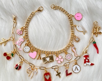 Op de afbeelding: Twee gouden bedelarmbanden met diverse kleurrijke bedels. Bedels zijn onder meer een roze hart, een rode chilipeper, een cowboylaars, een smiley, een bloem en letters. De armbanden liggen op een witte, zachte ondergrond.