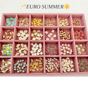 May include: A pink velvet jewelry tray with twenty-four compartments filled with assorted gold-tone metal charms. The charms feature various shapes including flowers, fruits, cameras, stars, sunglasses, and butterflies. Text reads EURO SUMMER.