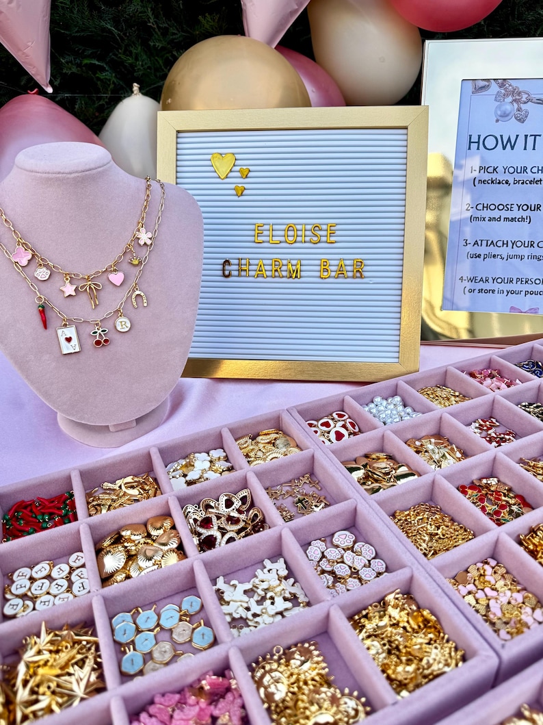 May include: A light pink velvet jewelry display bust wearing two gold-tone chain necklaces with various colorful charms, including a red chili, a playing card, cherries, and hearts, next to a letter board reading ELOISE CHARM BAR.