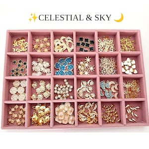 May include: A pink velvet jewelry tray with twenty compartments containing various metal charms. The charms feature celestial motifs including suns, moons, stars, planets, and lightning bolts in gold, white, blue, and black enamel finishes.