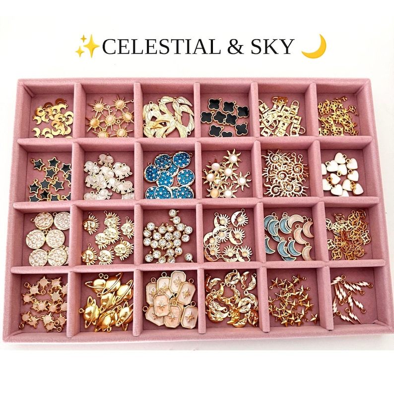 May include: A pink velvet jewelry tray with twenty compartments containing various metal charms. The charms feature celestial motifs including suns, moons, stars, planets, and lightning bolts in gold, white, blue, and black enamel finishes.