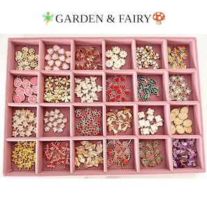 May include: A pink velvet jewelry tray with twenty compartments containing various metal charms. The charms feature designs including hearts, flowers, butterflies, strawberries, mushrooms, leaves, dragonflies, and roses. Text reads GARDEN & FAIRY.