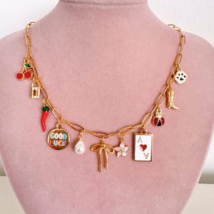 May include: A gold-toned chain necklace featuring various charms including red cherries, a gold padlock, a red chili pepper, a round disc with the text GOOD LUCK, a white pearl, a gold bow, a pink flower, an ace of hearts card, a ladybug, a boot, and a paw print.