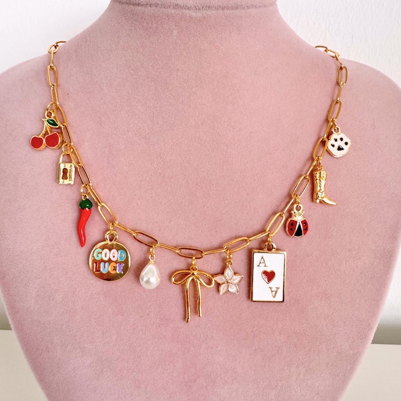 May include: A gold-toned chain necklace featuring various charms including red cherries, a gold padlock, a red chili pepper, a round disc with the text GOOD LUCK, a white pearl, a gold bow, a pink flower, an ace of hearts card, a ladybug, a boot, and a paw print.