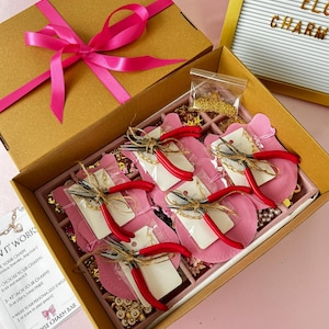 May include: A jewelry making kit in a brown cardboard box with a bright pink ribbon. The kit contains five pairs of pliers with red handles, small bags of gold-tone chains, various charms, and a printed instruction card labeled ELOISE CHARM BAR.