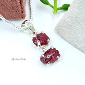 May include: A silver pendant necklace featuring two raw, deep red ruby gemstones. The rubies are set in a silver setting and hang from a delicate silver chain. The necklace is displayed on a white surface with a soft pink fabric in the background.